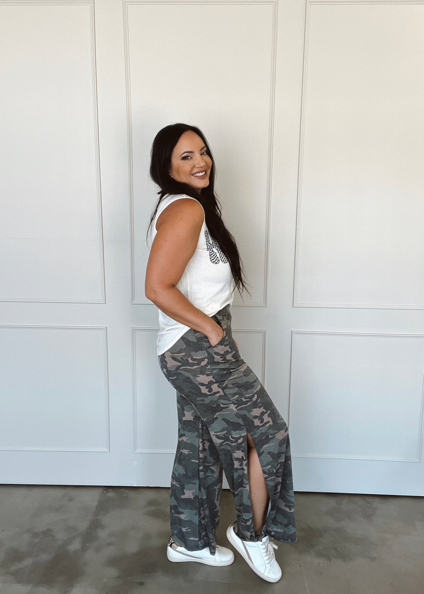 Woman wearing a white sleeveless top and camouflage pants standing against a white wall.