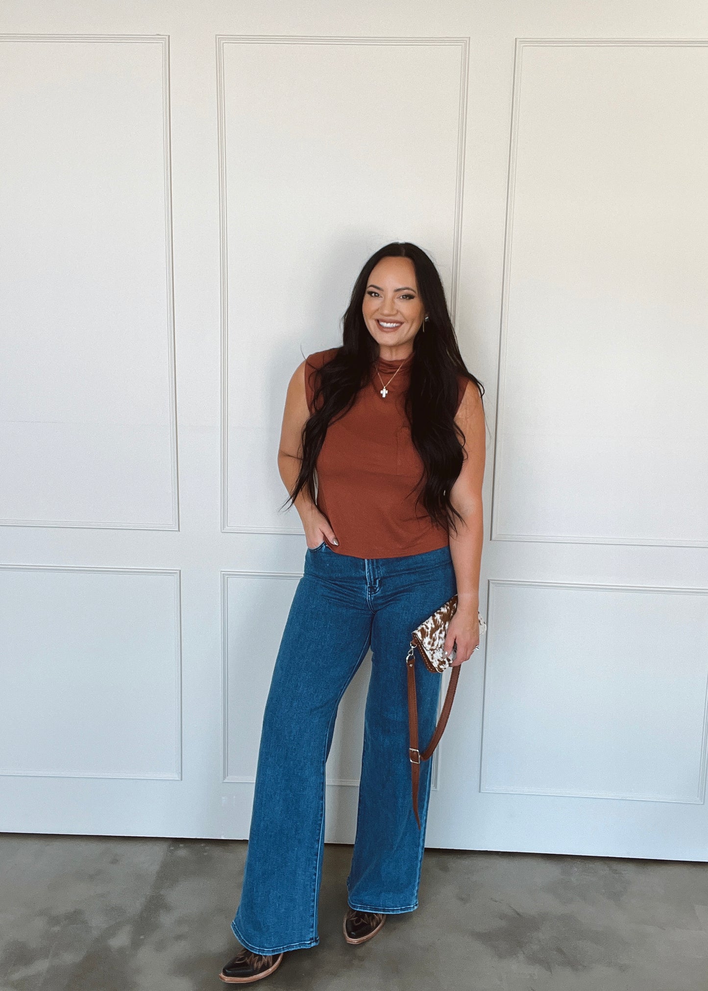 Woman wearing a brown top and blue jeans standing against a white wall.