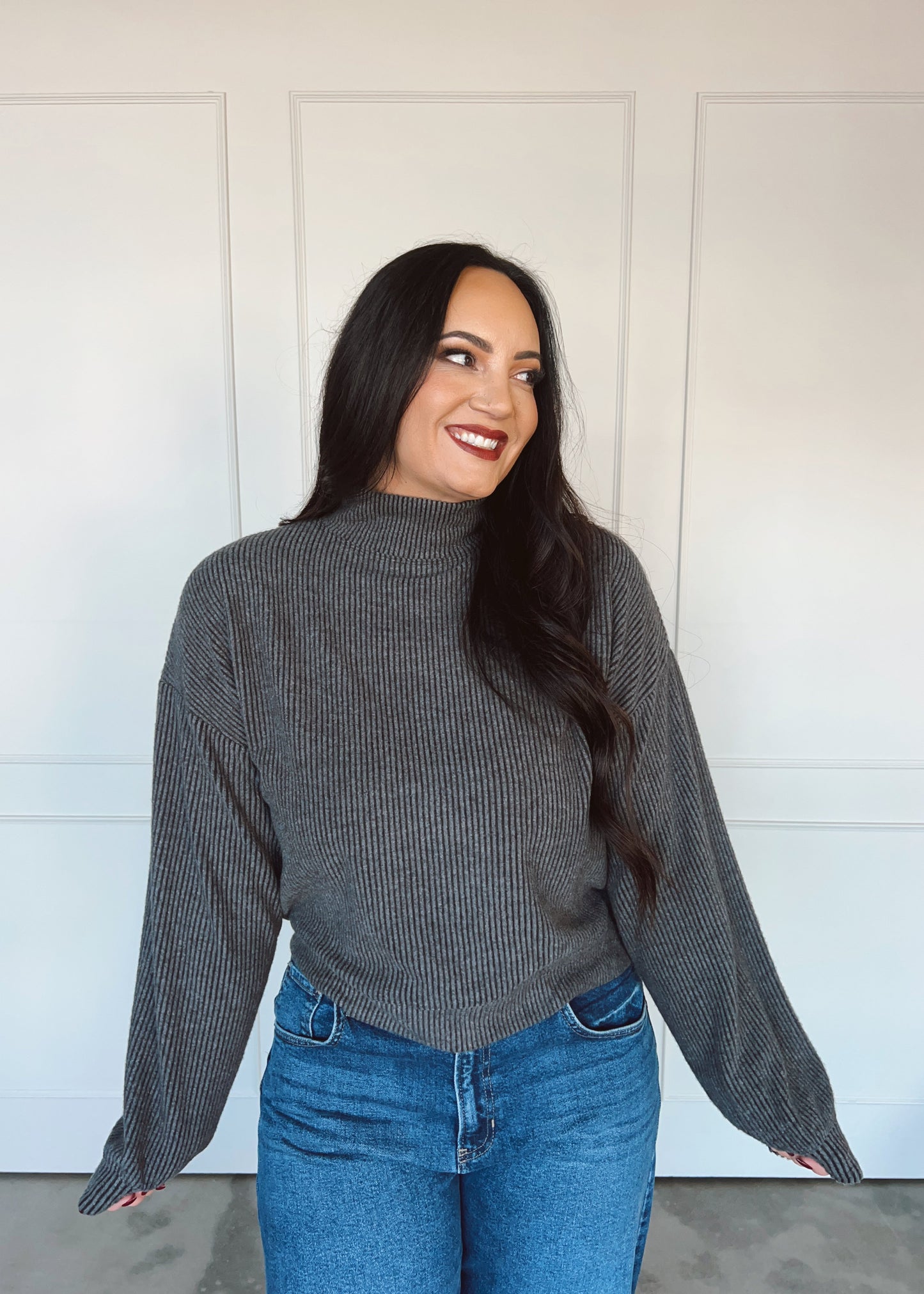 Woman wearing a gray ribbed sweater and blue jeans standing against a white wall.