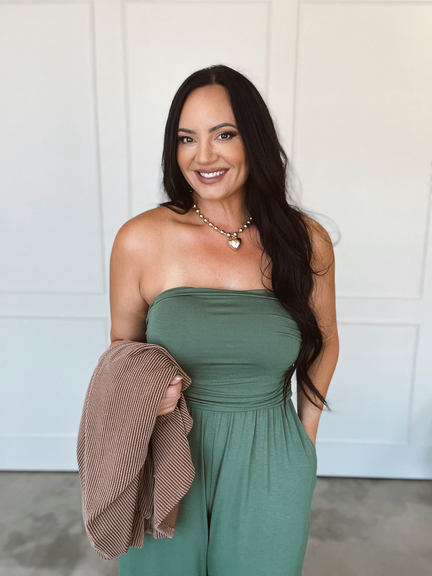 Woman wearing a green strapless jumpsuit holding a brown jacket against a white wall.
