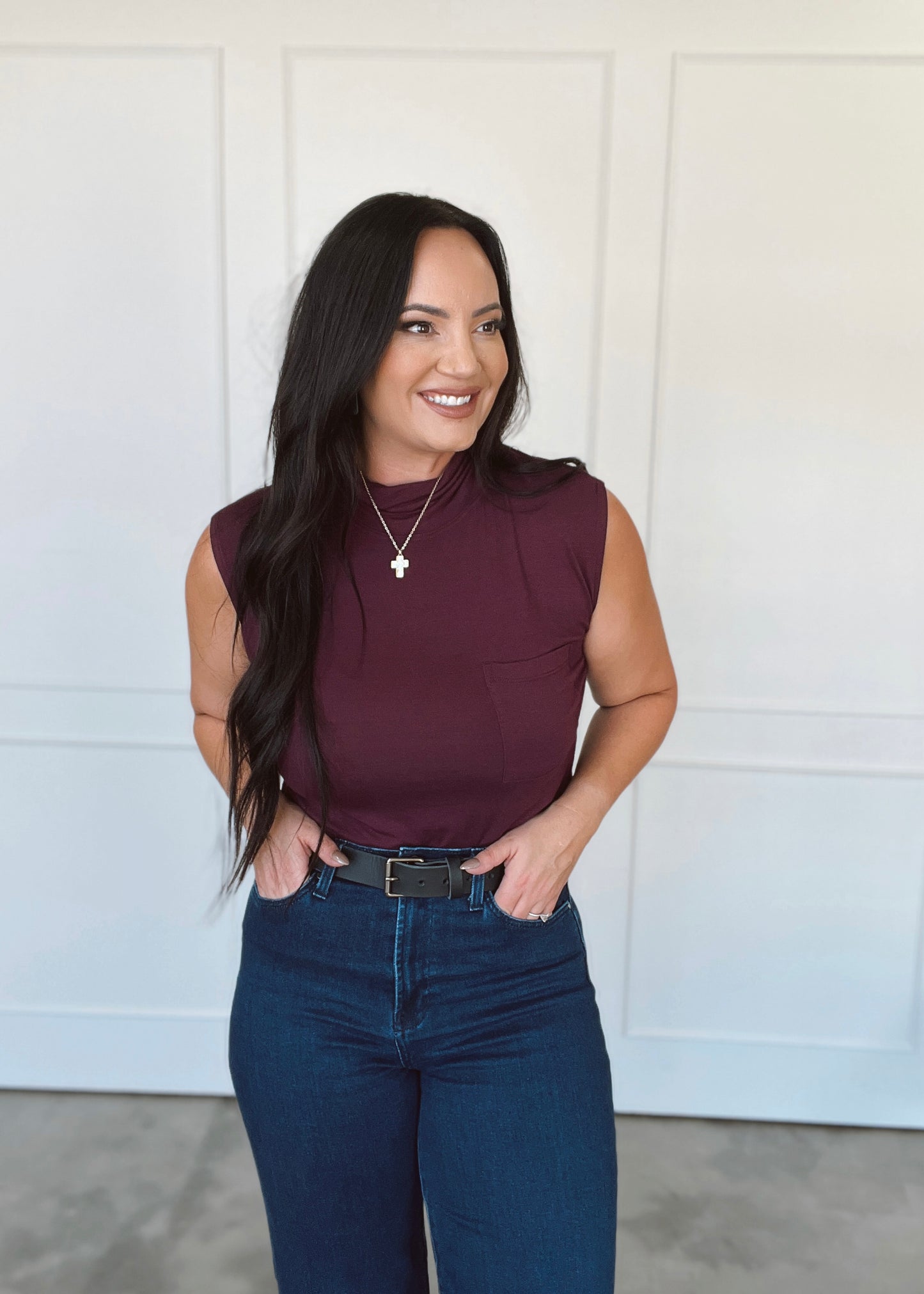 Woman wearing a maroon sleeveless top and blue jeans standing against a white wall.