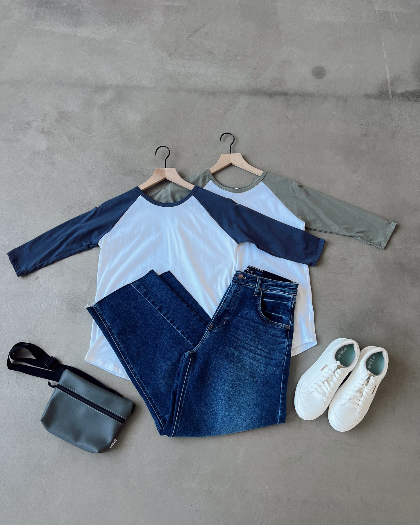 Fashionable outfit with a baseball shirt, jeans, sneakers, and a bag on a concrete floor.