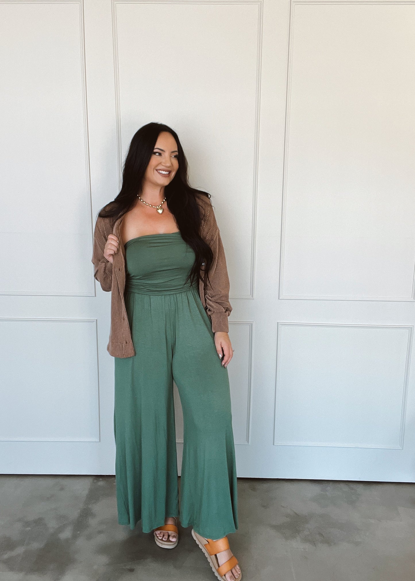 Woman wearing a green jumpsuit with a brown cardigan in front of a white paneled wall.