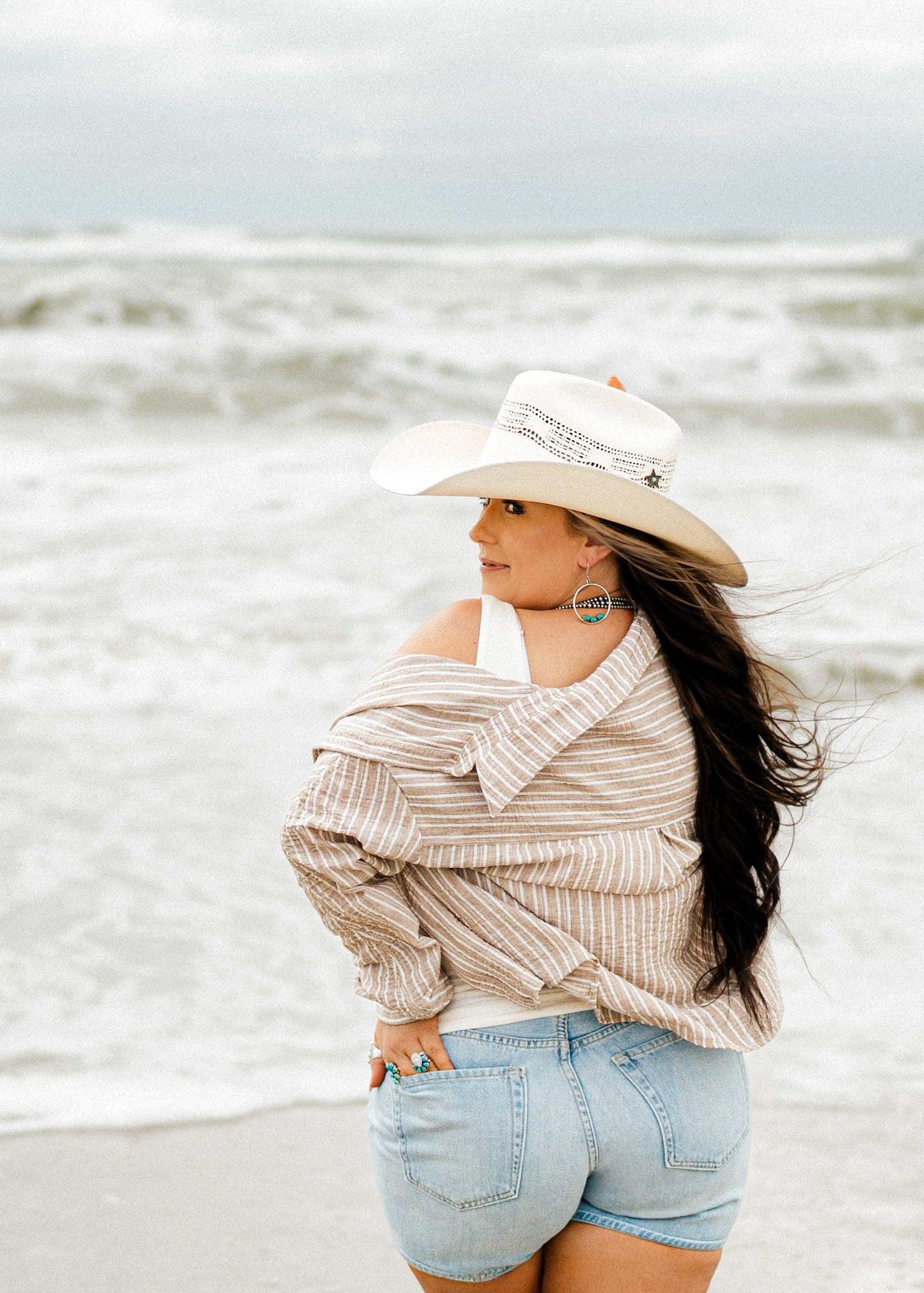 A woman stands on a beach facing the ocean, wearing a cream wide-brim hat, a beige and white striped off-the-shoulder shirt, and light blue denim shorts.
