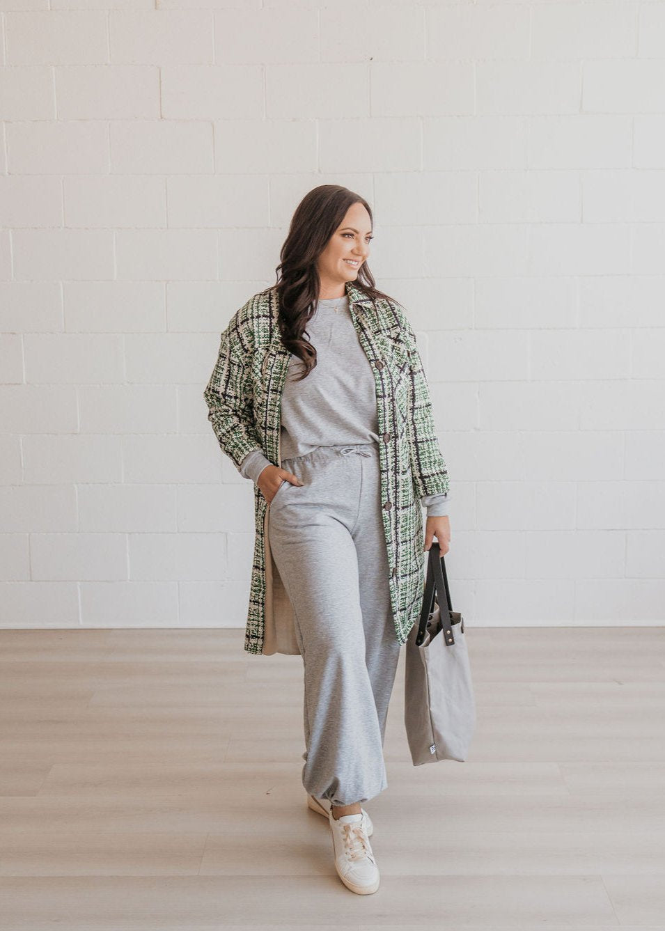 A woman wearing a green and gray checkered coat, gray pants with pockets, and white sneakers, standing in a studio setting.