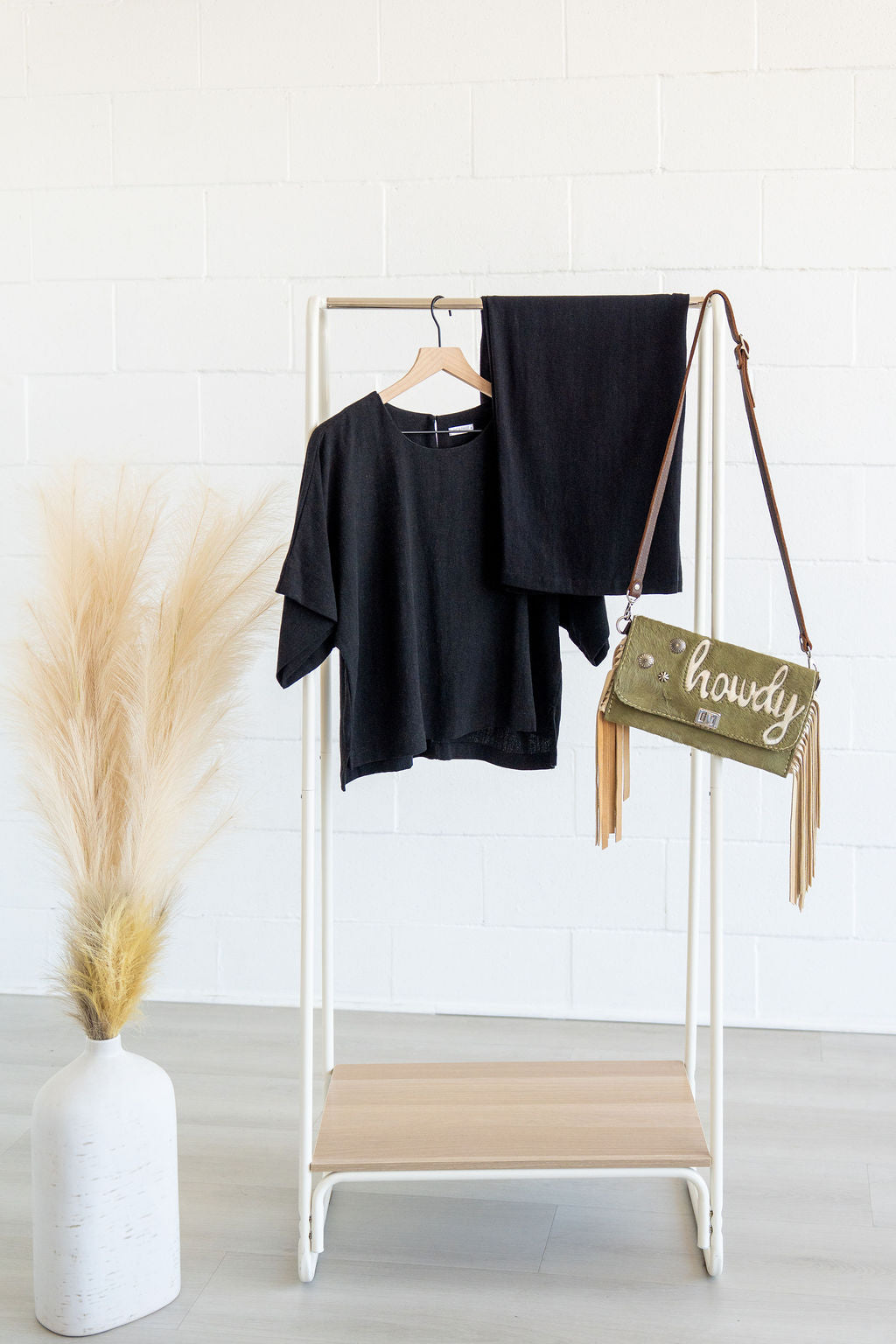 Clothing rack with black shirt, black pants, and green bag labeled 'howdy' against a white wall.