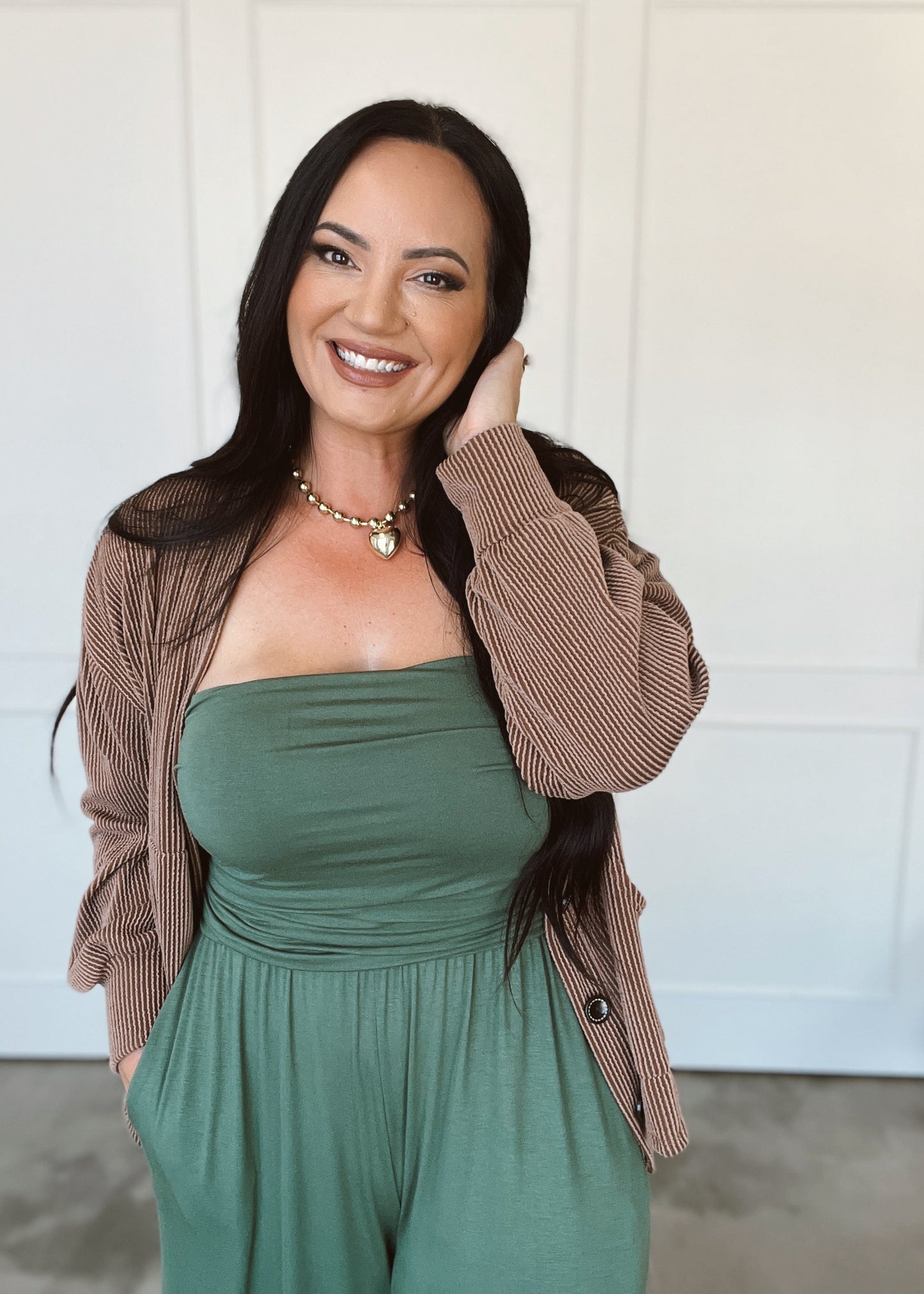 Woman wearing a green strapless jumpsuit with a brown cardigan against a white background