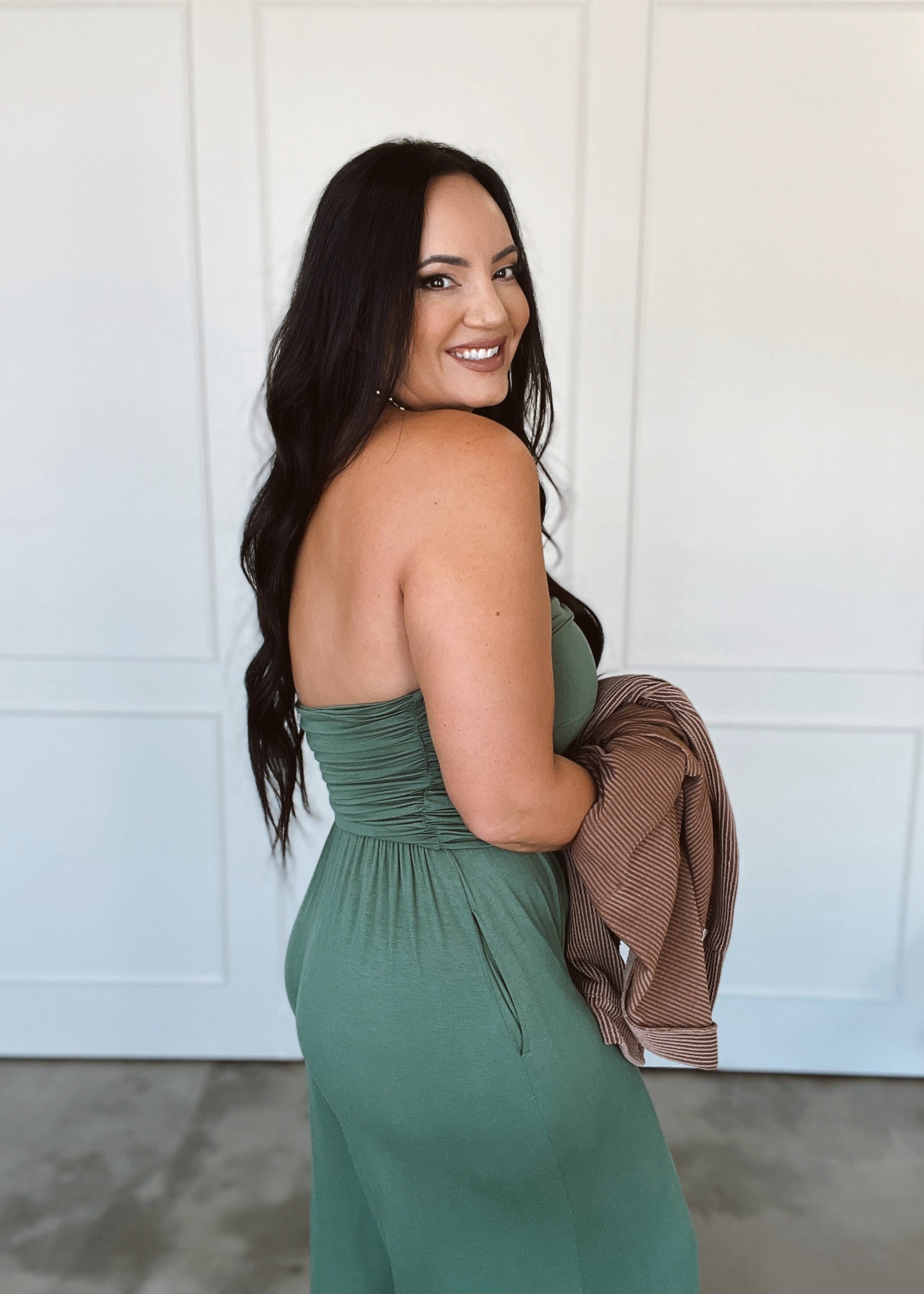 Woman in a green dress holding a brown jacket against a white wall.