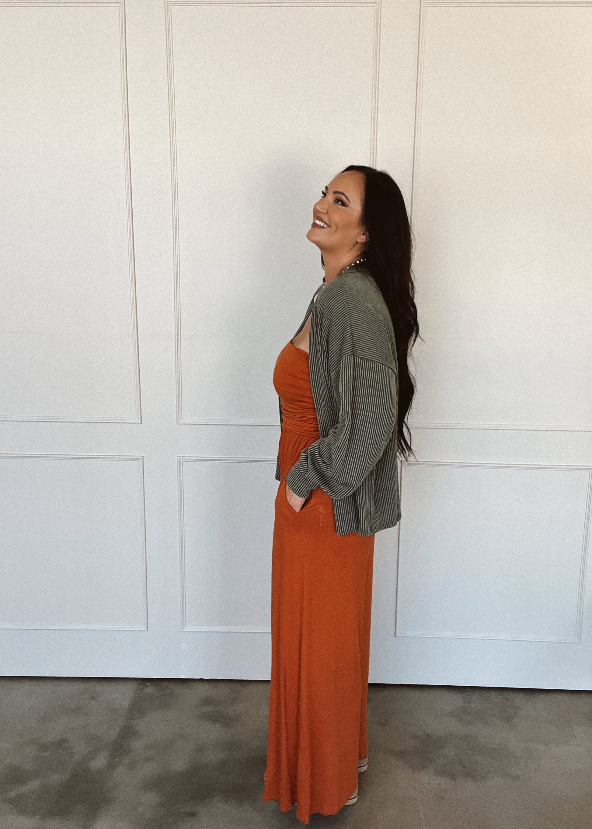 Woman wearing a rust-colored dress and green cardigan standing against a white paneled wall.