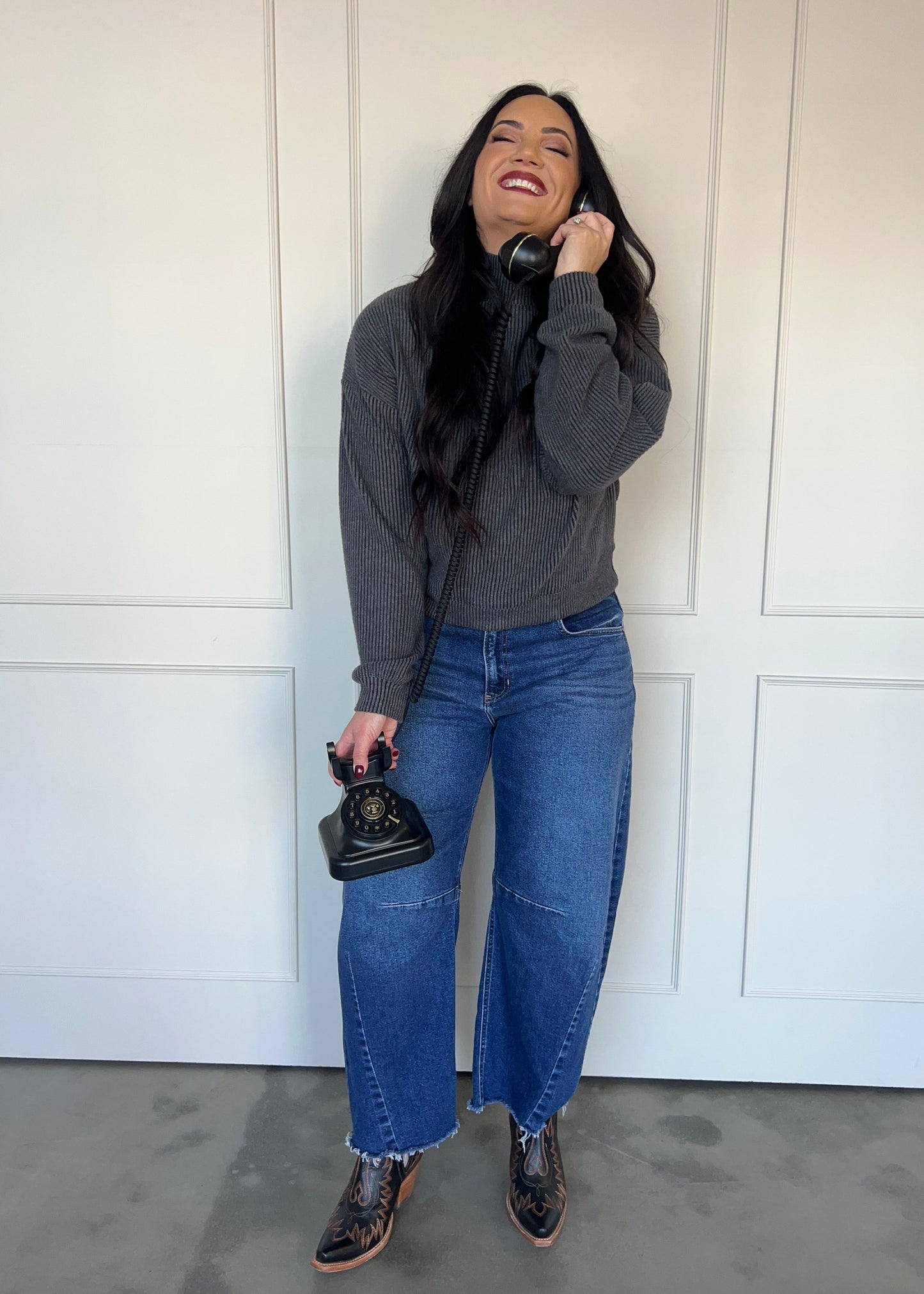 Woman wearing a gray sweater and blue jeans, holding a vintage phone against a white paneled wall.
