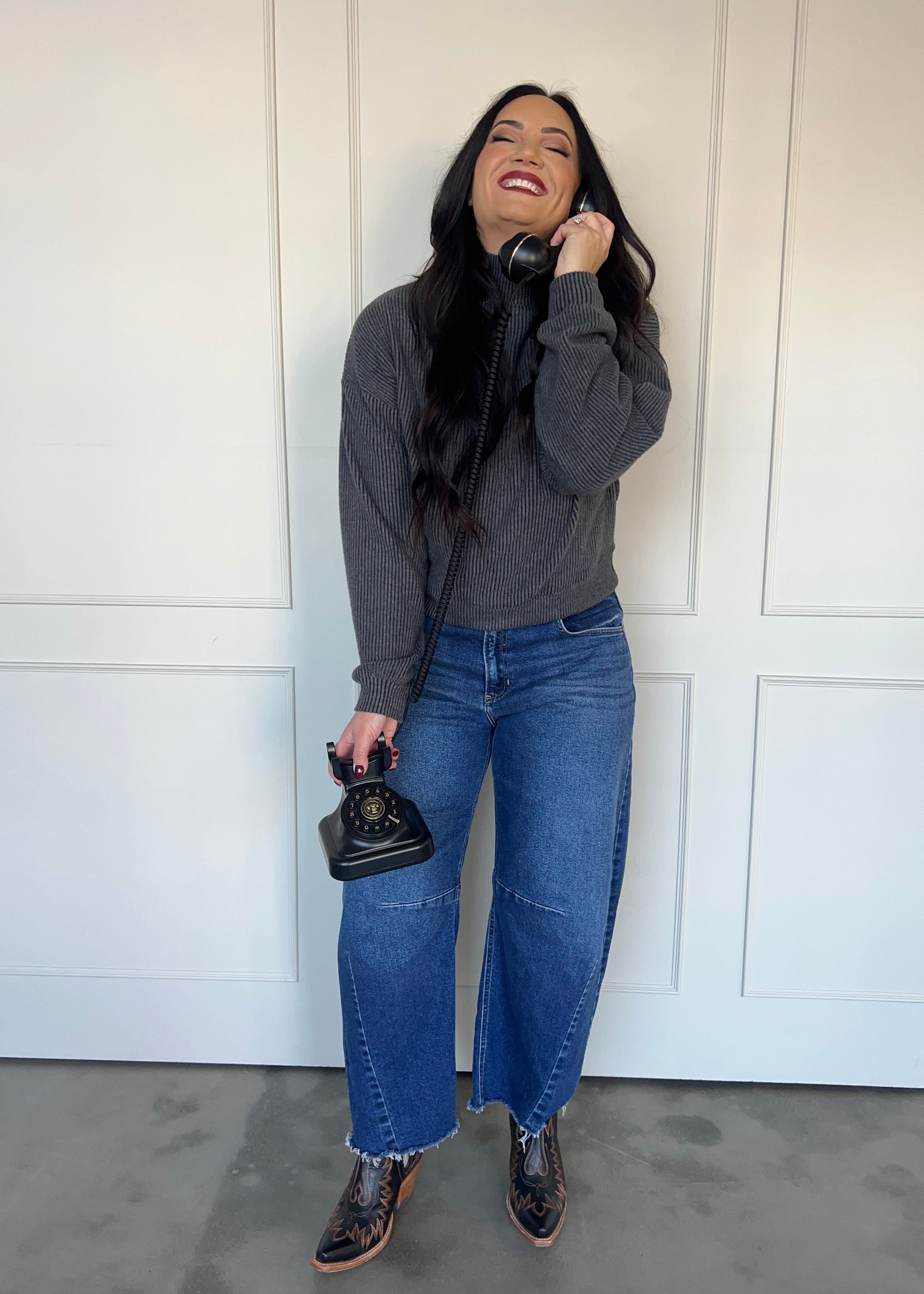 Woman wearing a gray sweater and blue jeans, holding a vintage phone against a white paneled wall.