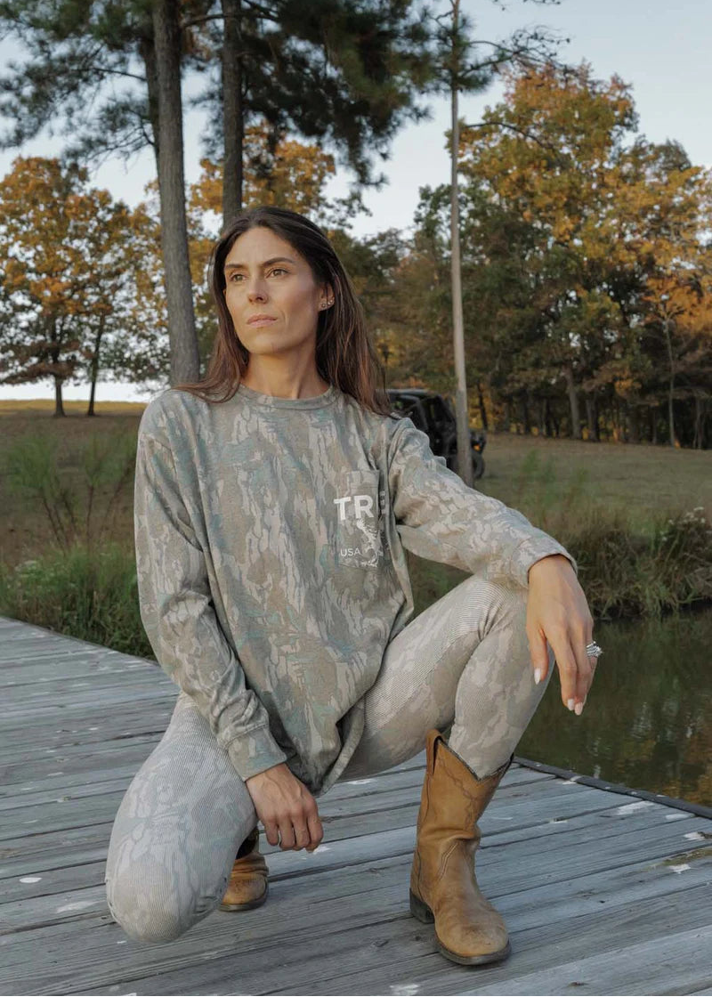 Woman in camouflage outfit squatting on a wooden dock with trees and water in the background