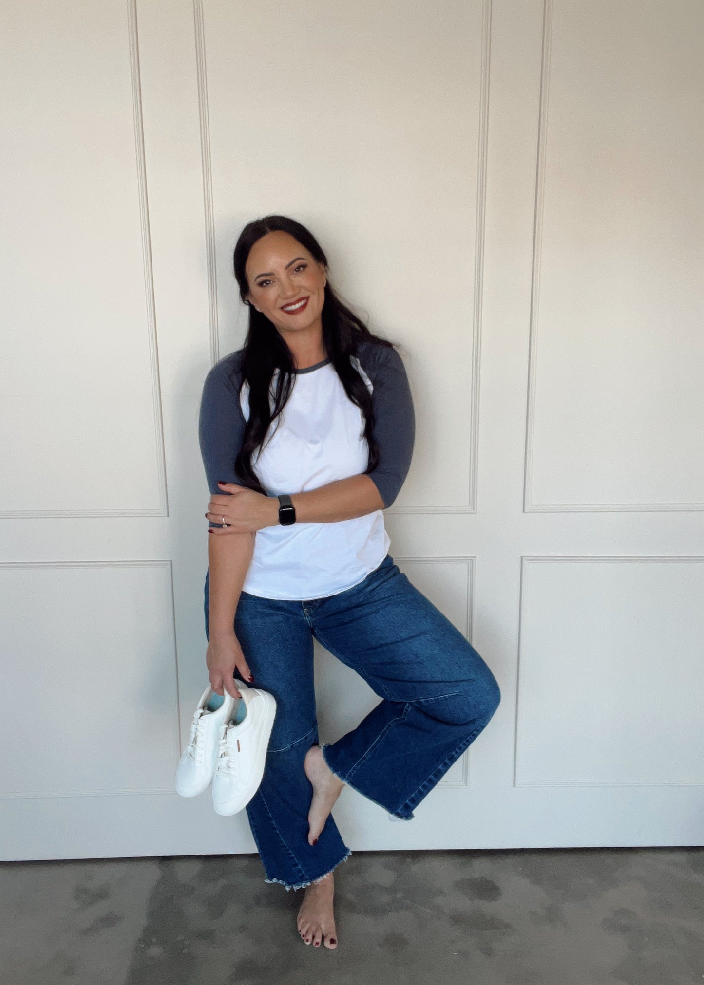 Woman in casual outfit holding white shoes against a white wall.