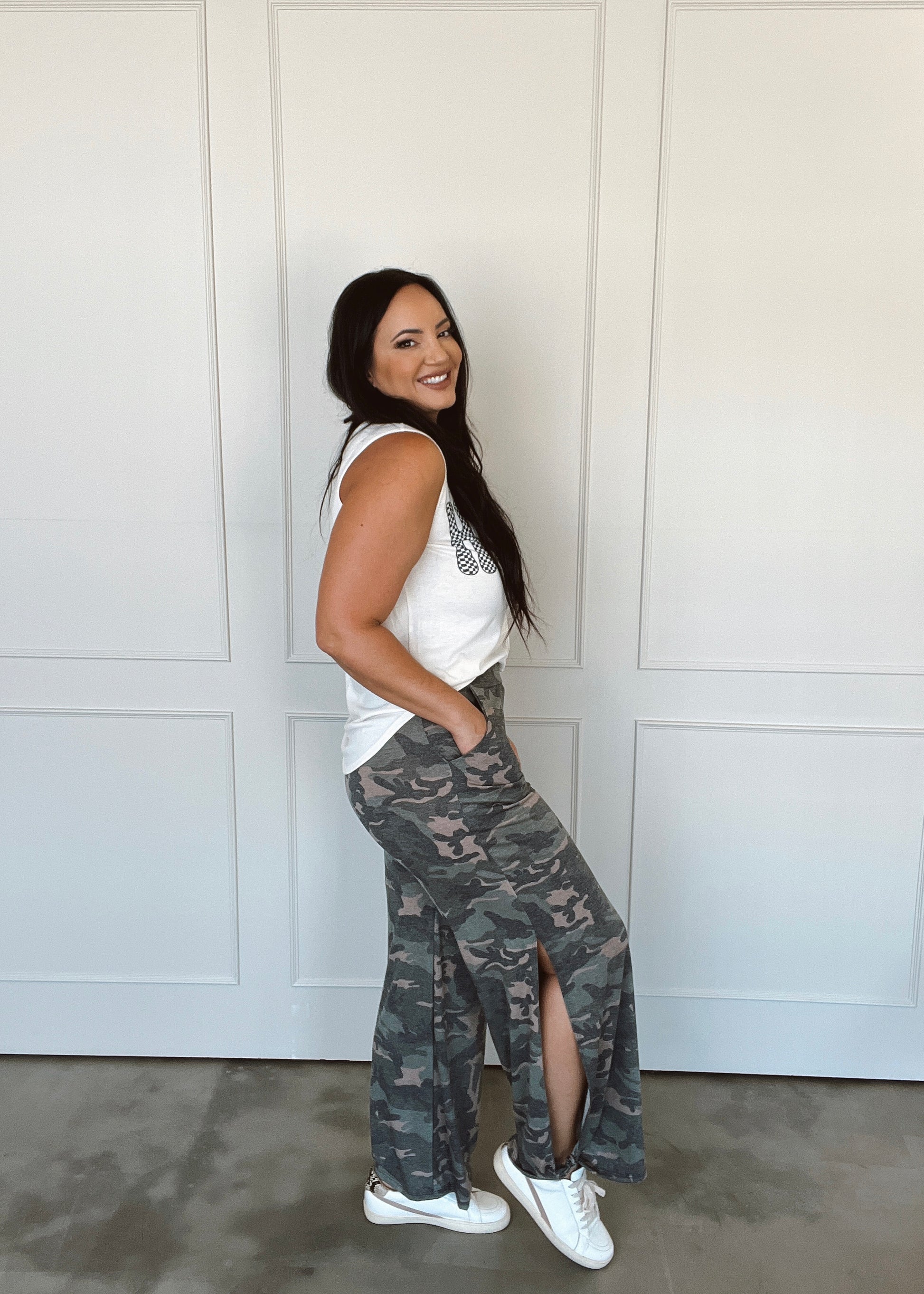 Woman wearing a white sleeveless top and camouflage pants standing against a white wall.