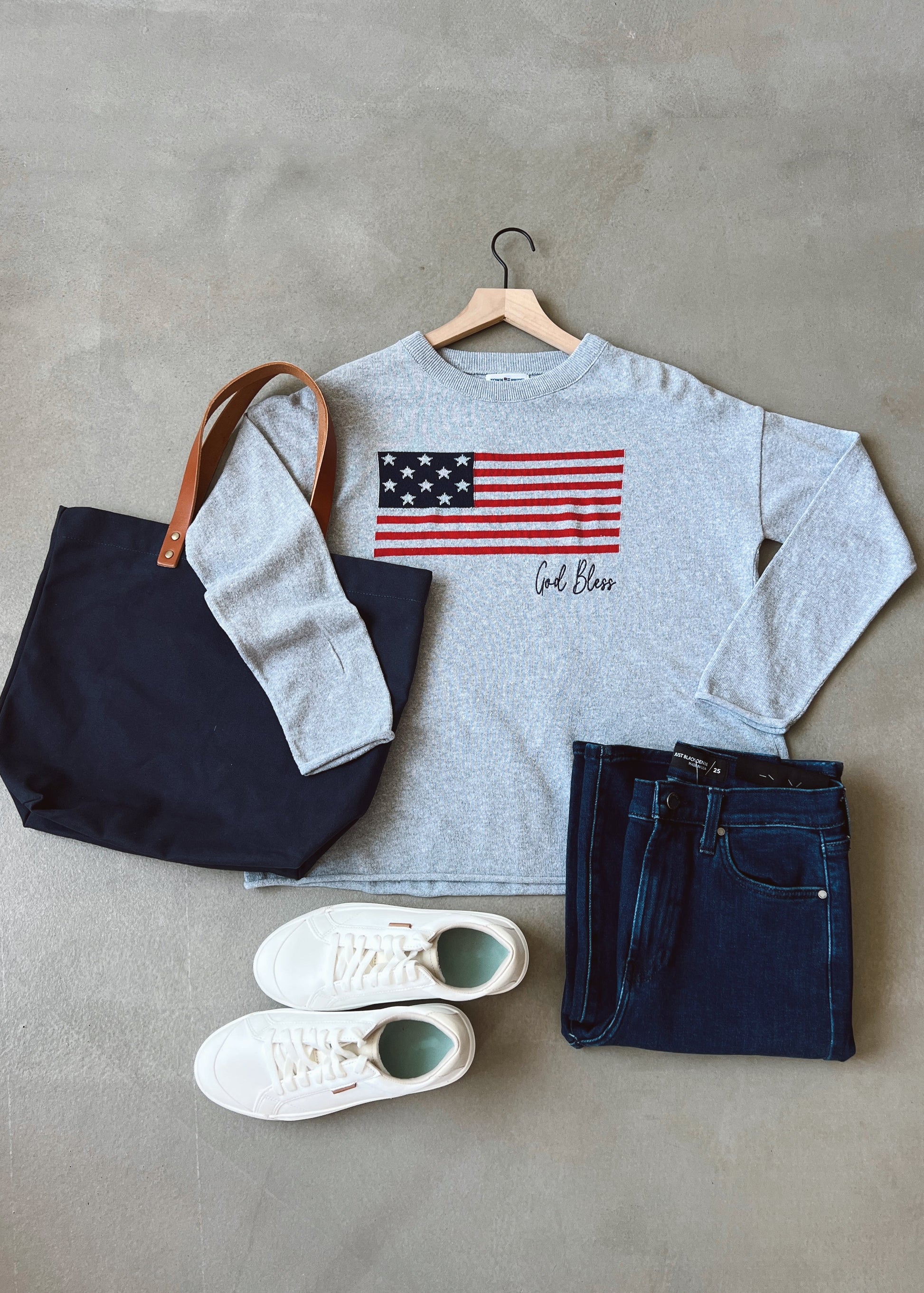 Gray sweatshirt with American flag design, navy tote bag, white sneakers, and dark jeans on a gray surface.