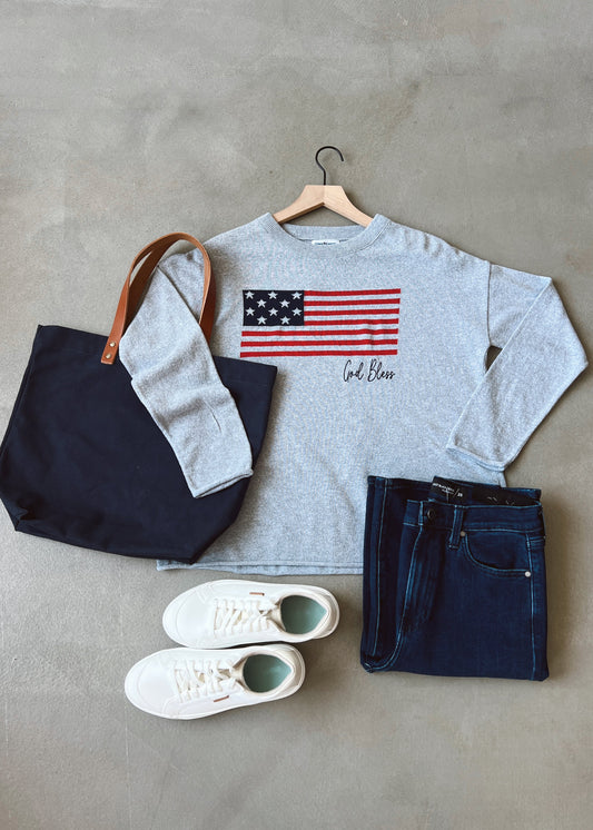 Gray sweatshirt with American flag design, navy tote bag, white sneakers, and dark jeans on a gray surface.