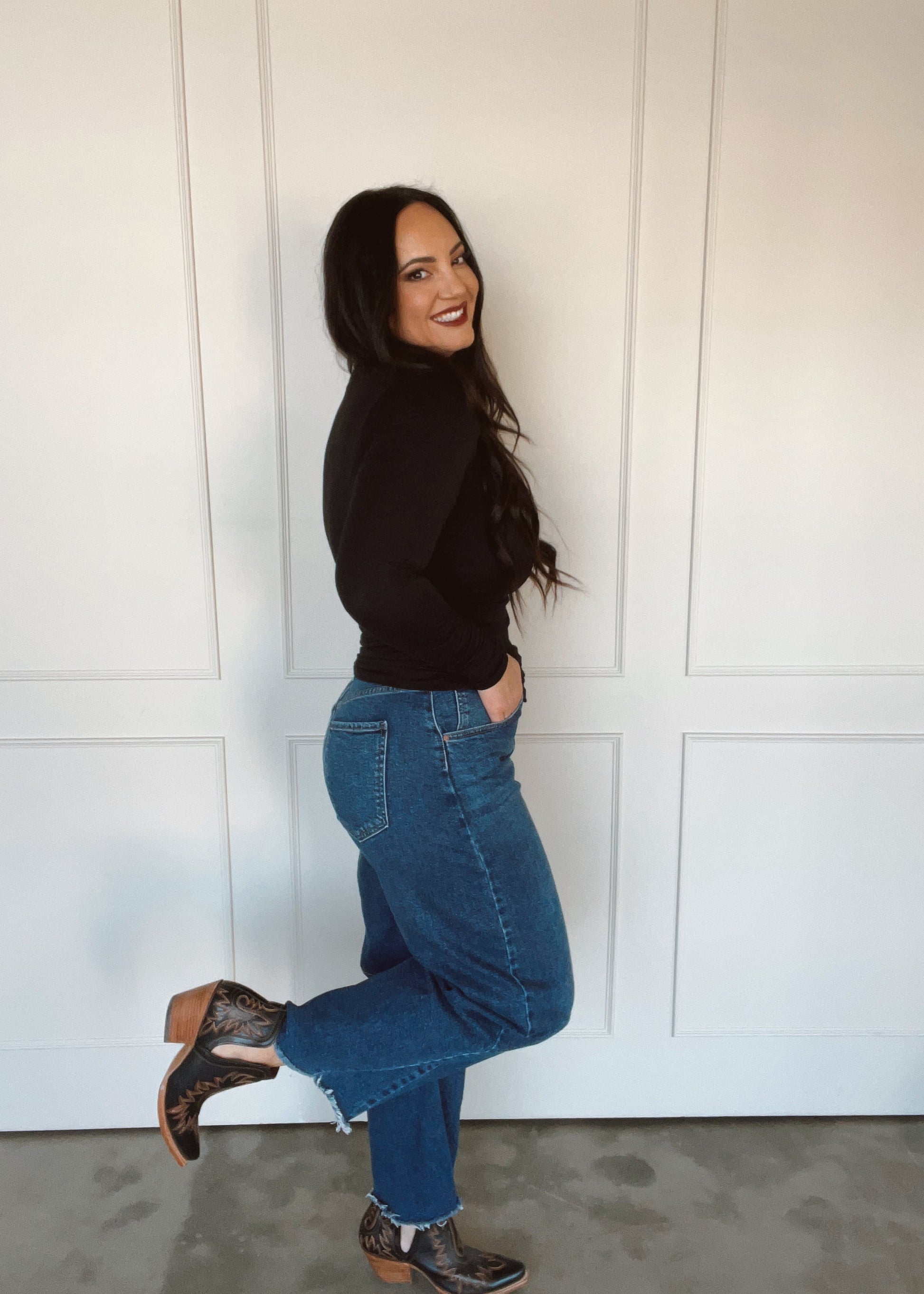 Woman wearing a black top and blue jeans standing against a white paneled wall.