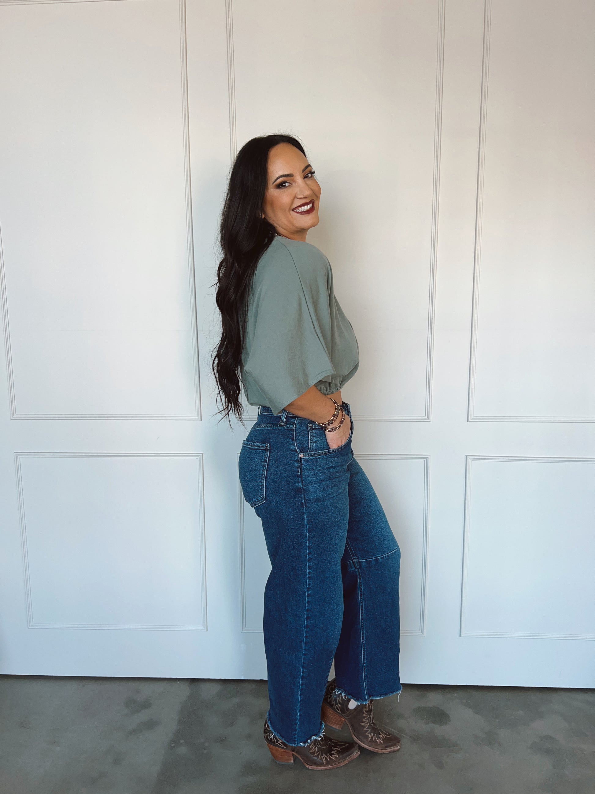 Woman wearing a green top and blue jeans standing against a white paneled wall.