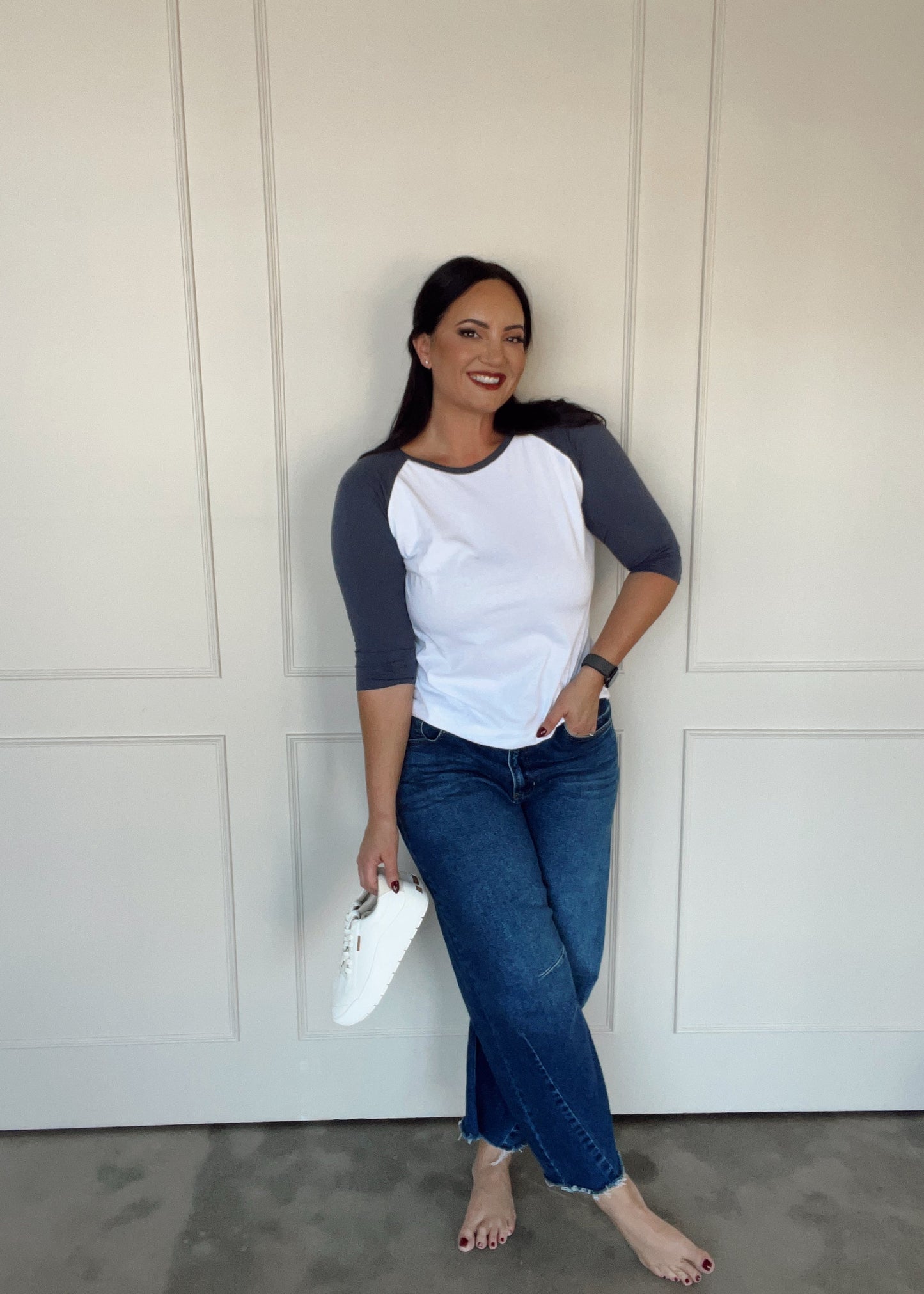 Woman wearing a white and gray baseball shirt with blue jeans, standing against a white paneled wall.
