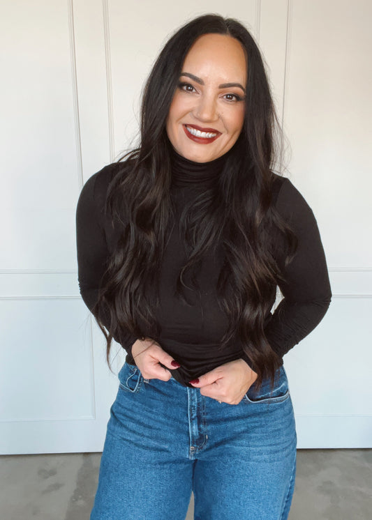Woman wearing a black long-sleeve top and blue jeans against a white wall.