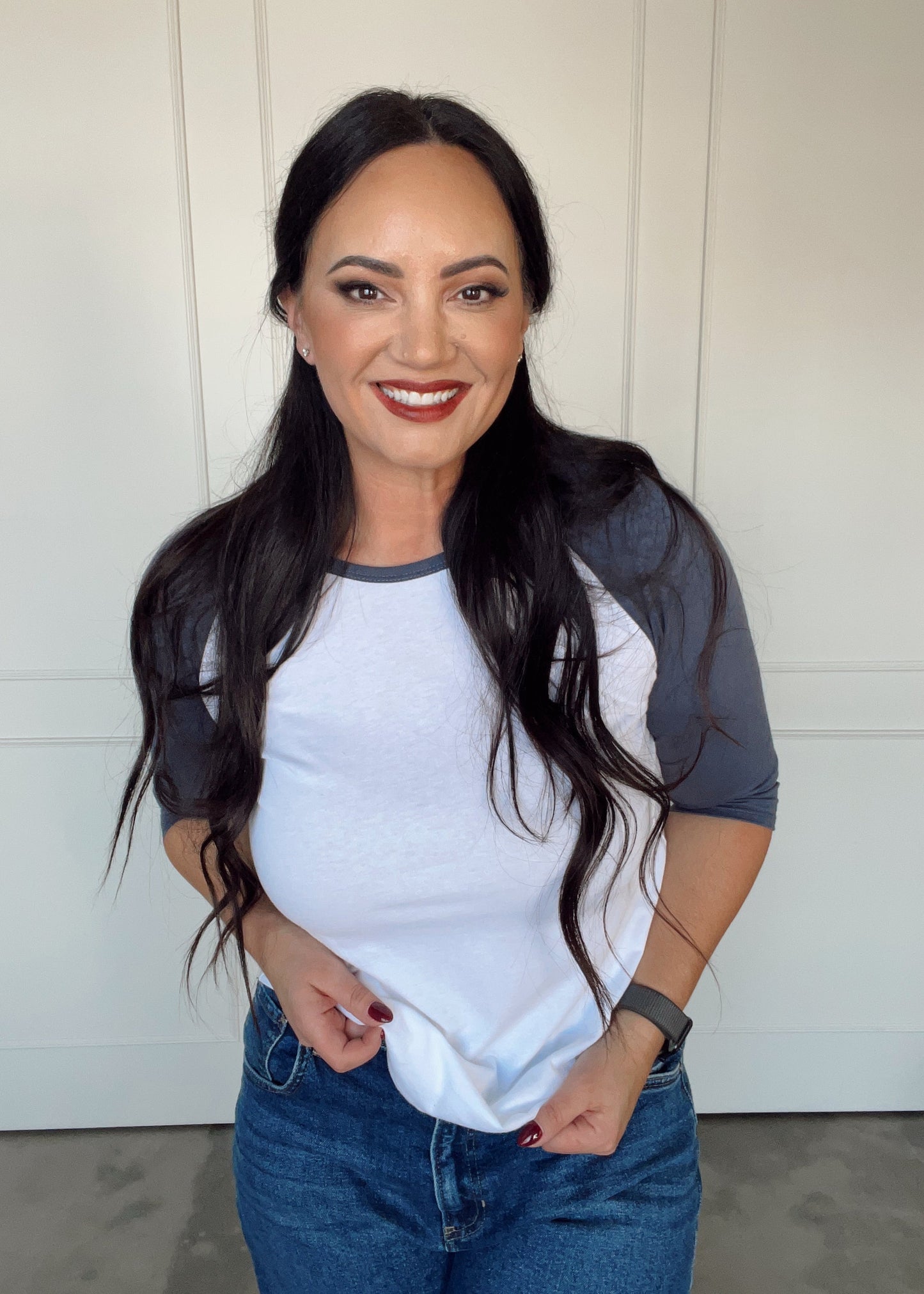 Woman wearing a white and gray baseball shirt with blue jeans against a white wall.