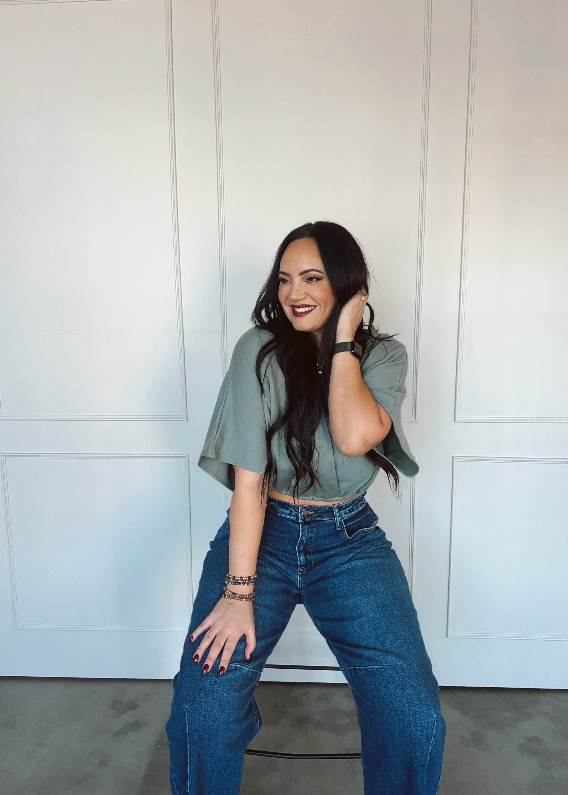Woman wearing a green top and blue jeans posing against a white paneled wall.