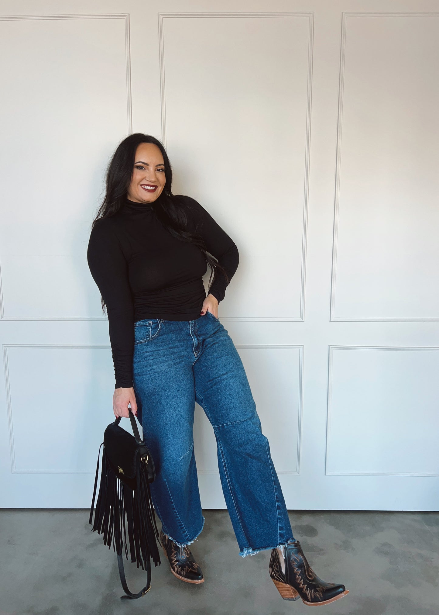 Woman wearing a black top, blue jeans, and brown boots against a white paneled wall.