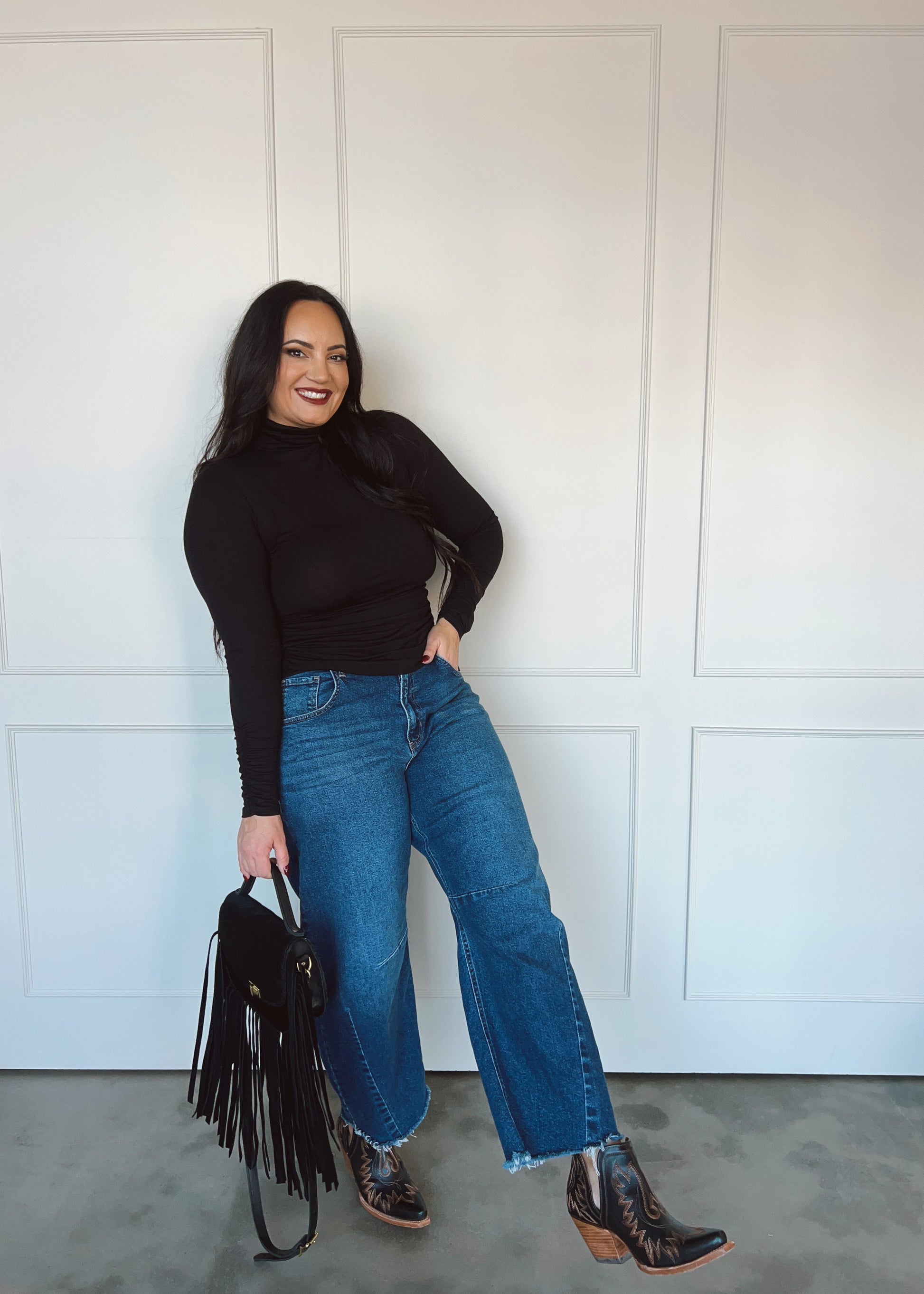 Woman wearing a black top, blue jeans, and brown boots against a white paneled wall.