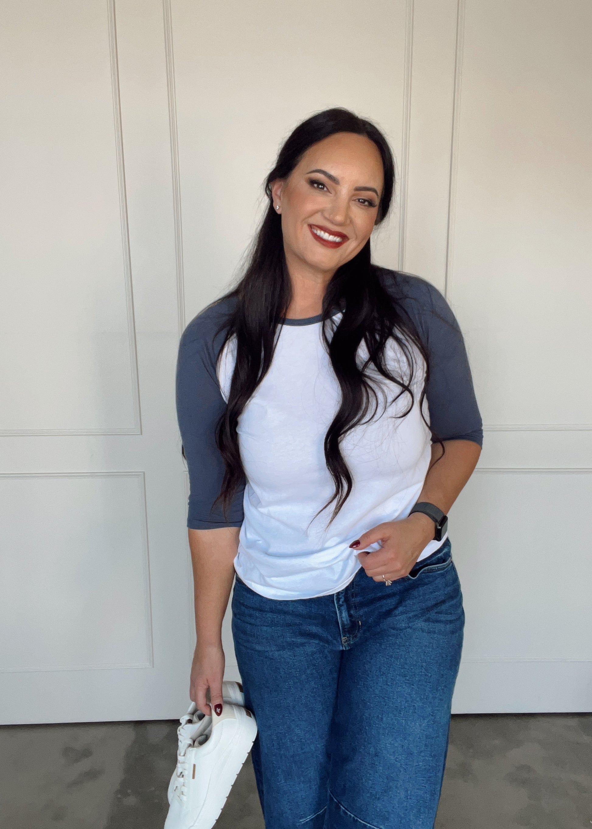 Woman wearing a white and blue baseball shirt with jeans standing in front of a white door.