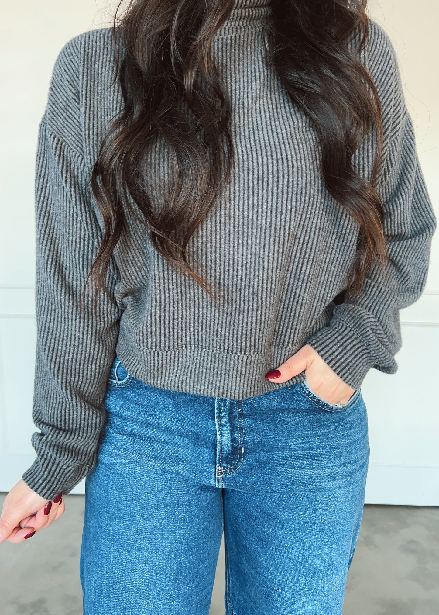 Person wearing a gray ribbed sweater and blue jeans with a plain background