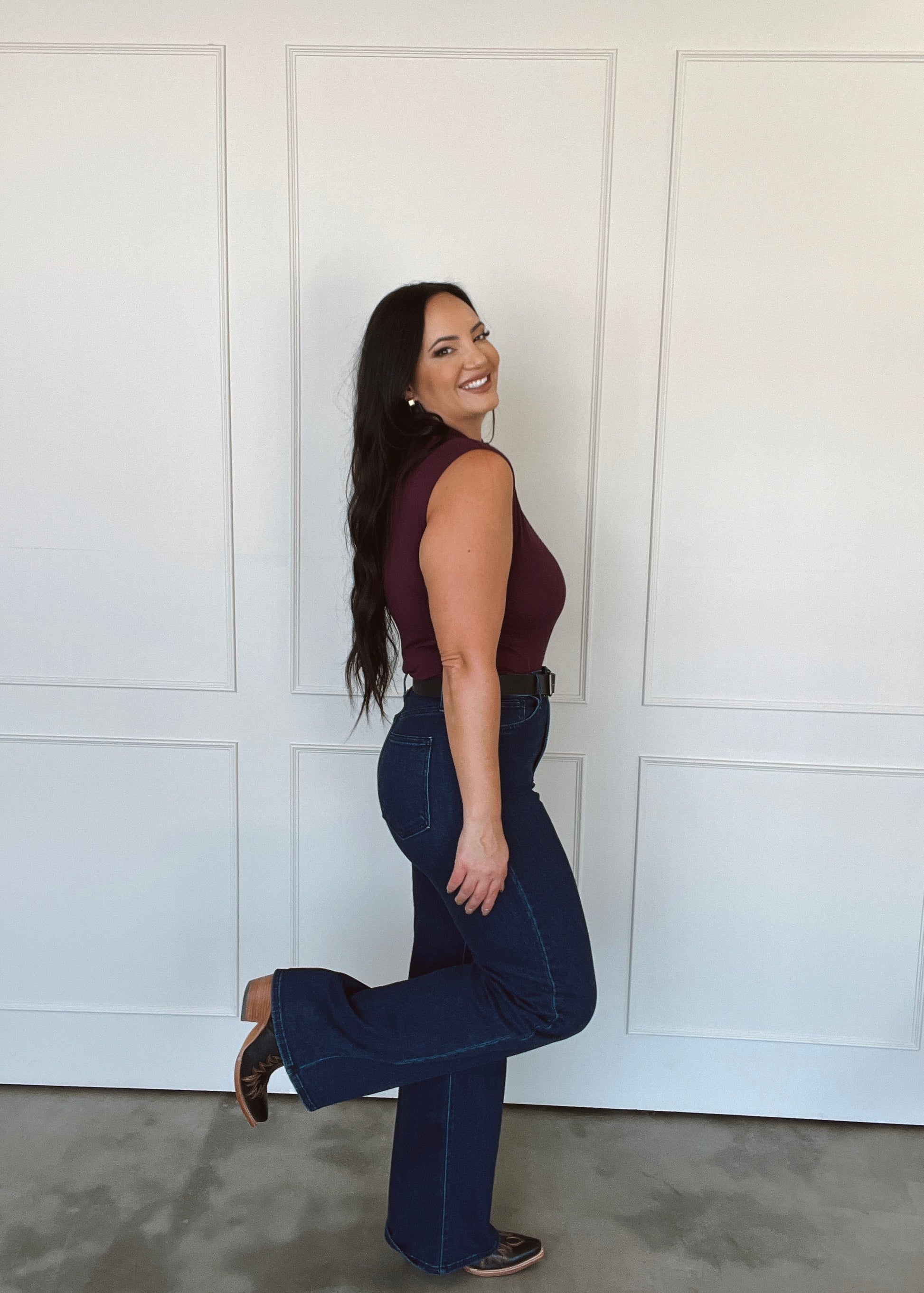 Woman wearing a maroon top and blue jeans posing against a white paneled wall.