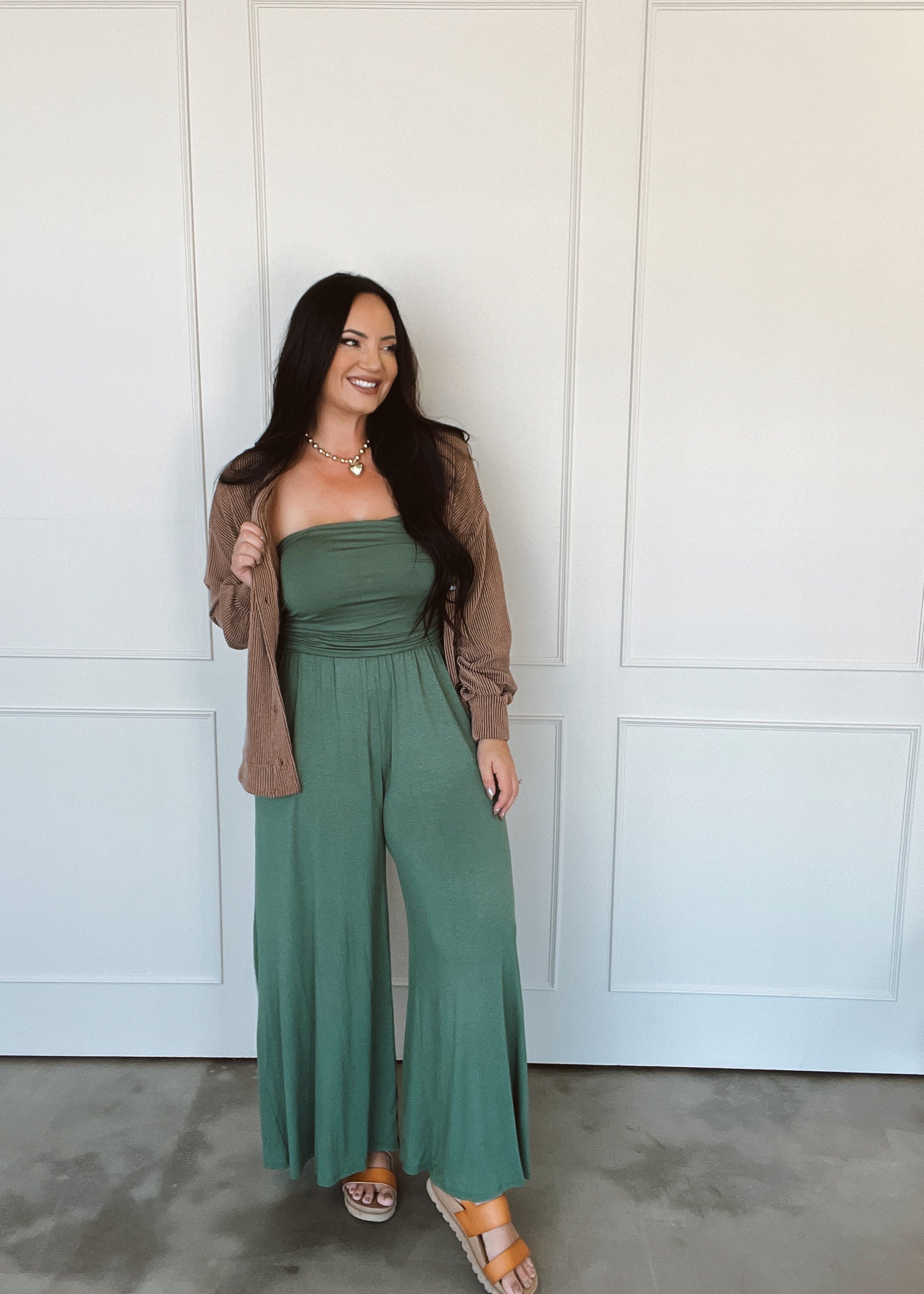 Woman wearing a green jumpsuit with a brown cardigan in front of a white paneled wall.