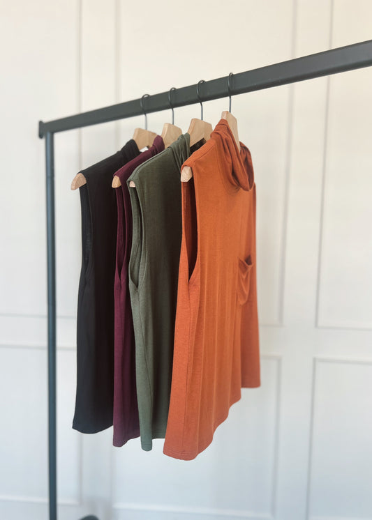 Four shirts in black, burgundy, green, and orange hanging on a rack against a white wall.
