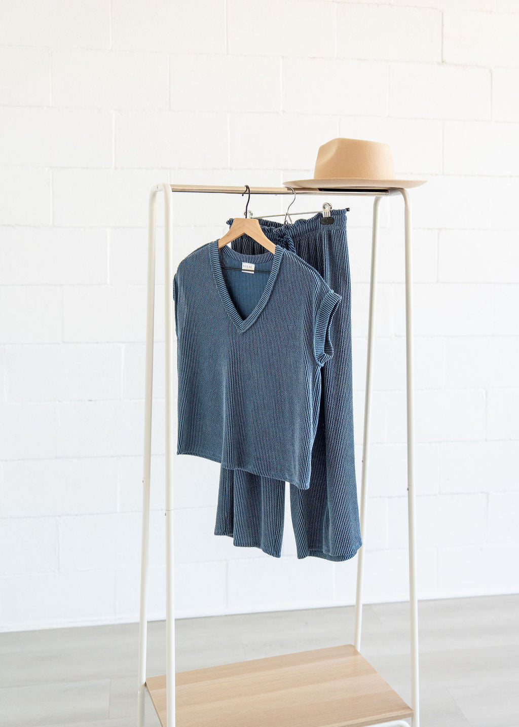 Blue sweater and pants on a rack with a beige hat against a white brick wall.